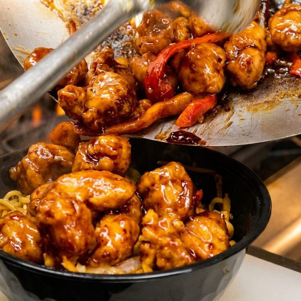Glazed orange chicken pieces and red peppers slide from a wok into a black bowl of noodles.