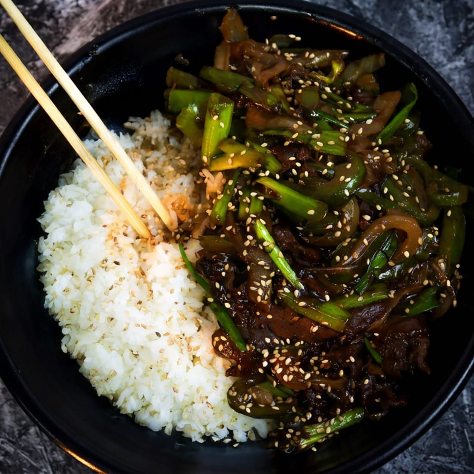 A black bowl of white rice and stir-fried beef with green peppers, onions, and sesame seeds.