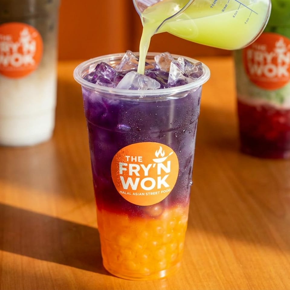 Green liquid pouring into a purple and orange layered drink with ice and boba, on a wooden table.
