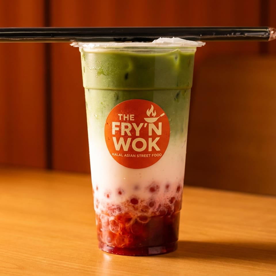 A Fry'N Wok branded layered drink with green matcha, white milk, and red boba on a wooden table.